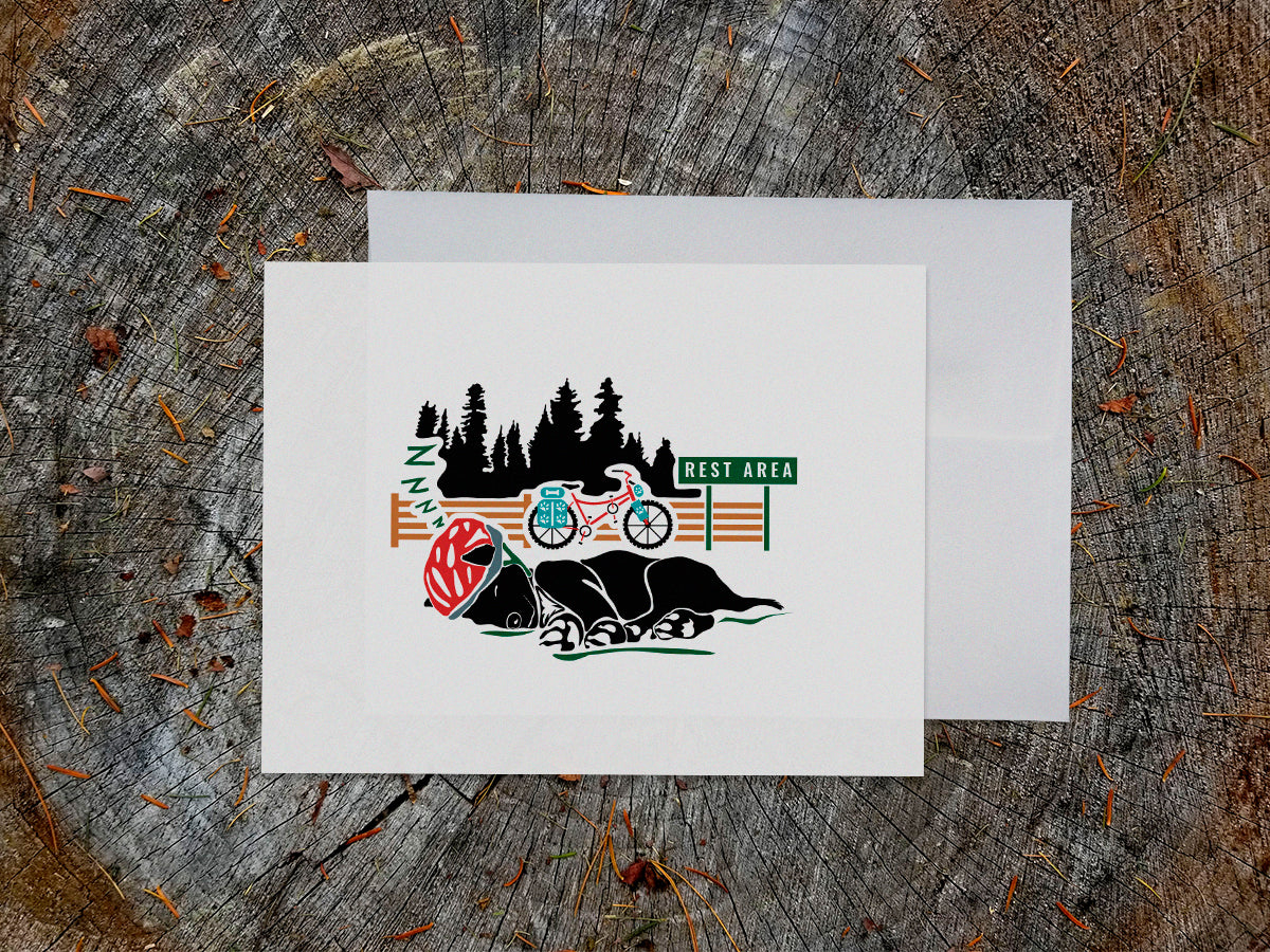 Greeting card of black dog with a red helmet sleeping with a bike and sign that reads Rest Area in the background