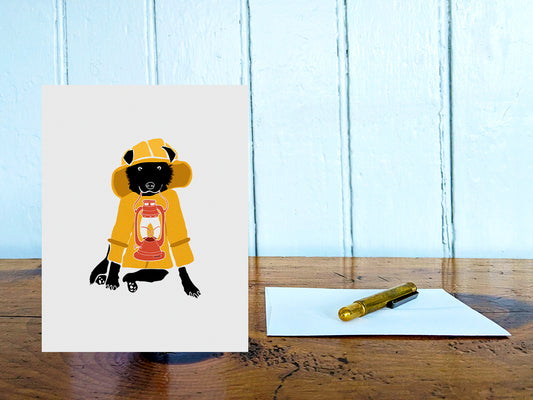 Greeting card with image of a dog with a yellow fisherman's jacket holding a lamp on a table with a pen and envelope.