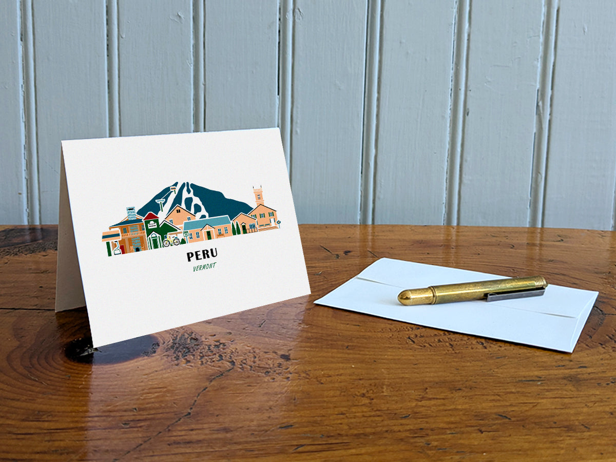 Greeting card with image of buildings arranged in a city skyline with the text Peru Vermont on a table with gold pen and envelope.