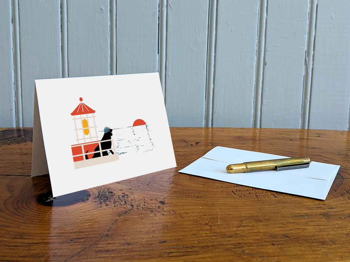 Greeting card with image of a dog in a lighthouse on a table with gold pen and envelope.