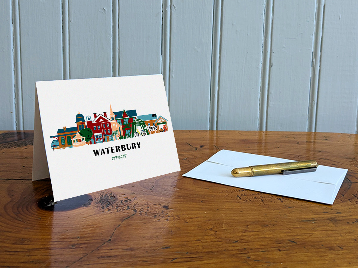 Greeting card with image of buildings arranged in a city skyline with the text Waterbury Vermont on a table with gold pen and envelope.