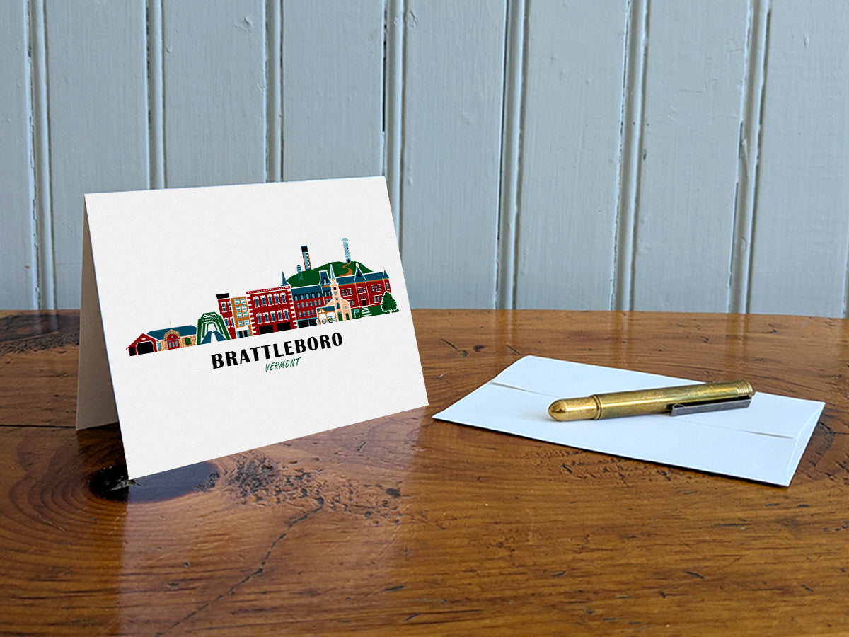Greeting card with image of buildings arranged in a city skyline with the text Brattleboro Vermont on a table with gold pen and envelope.
