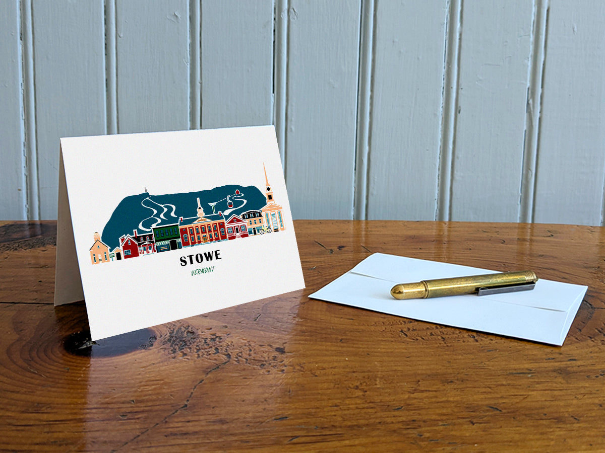 Greeting card with image of buildings arranged in a city skyline with the text Stowe Vermont on a table with gold pen and envelope.