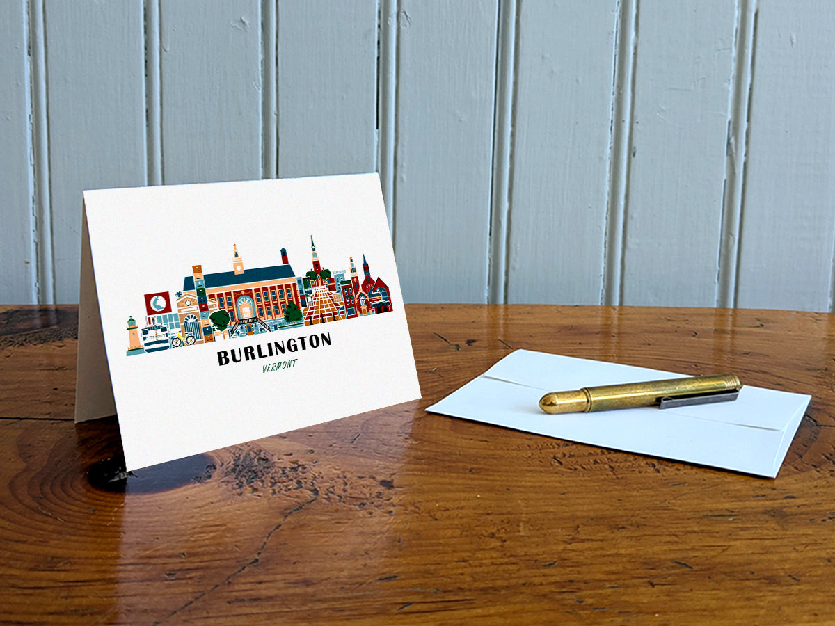 Greeting card with image of buildings arranged in a city skyline with the text Burlington Vermont on a table with gold pen and envelope.