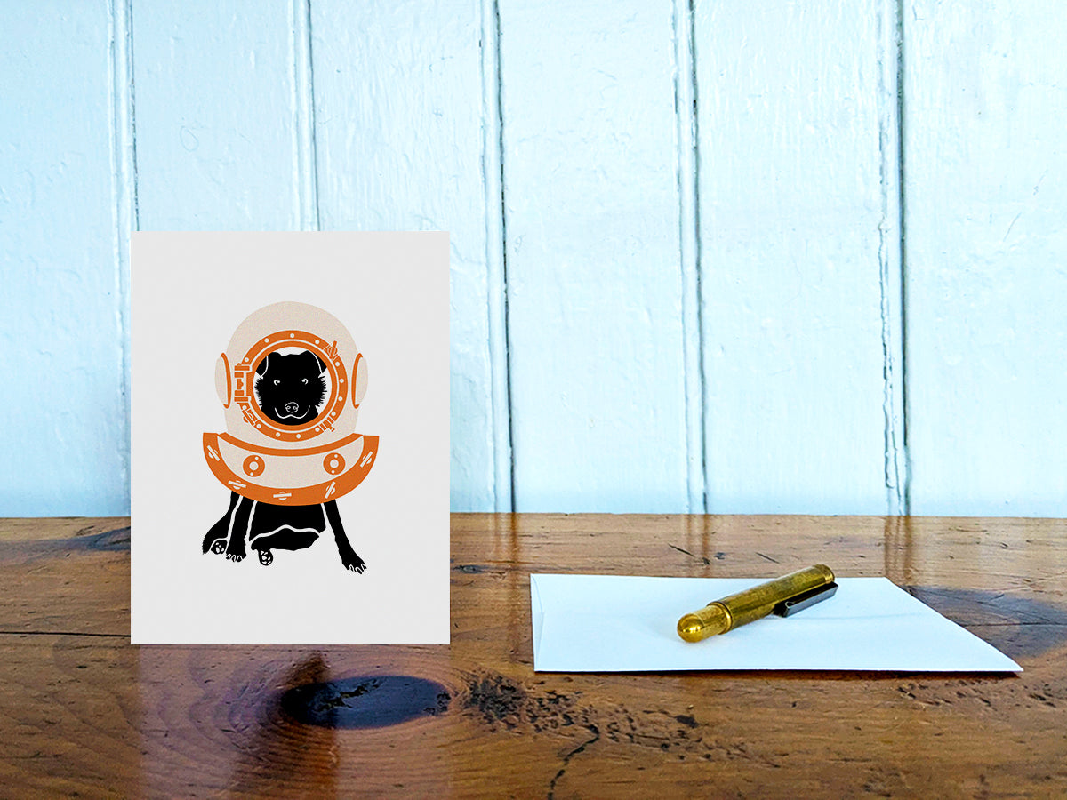 Greeting card with image of a dog with a scuba diving helmet on a table with gold pen and envelope.