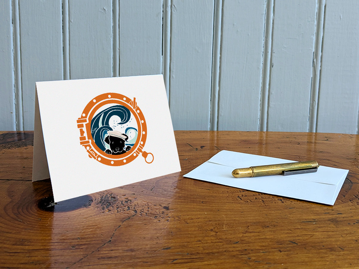Greeting card with image of a dog looking through a porthole on a table with gold pen and envelope.