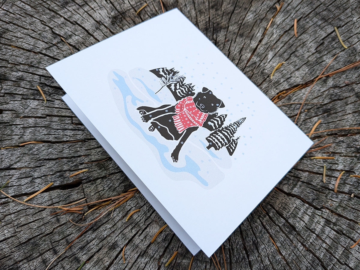 Greeting card with image of black dog with red scarf among a snowy winter landscape.