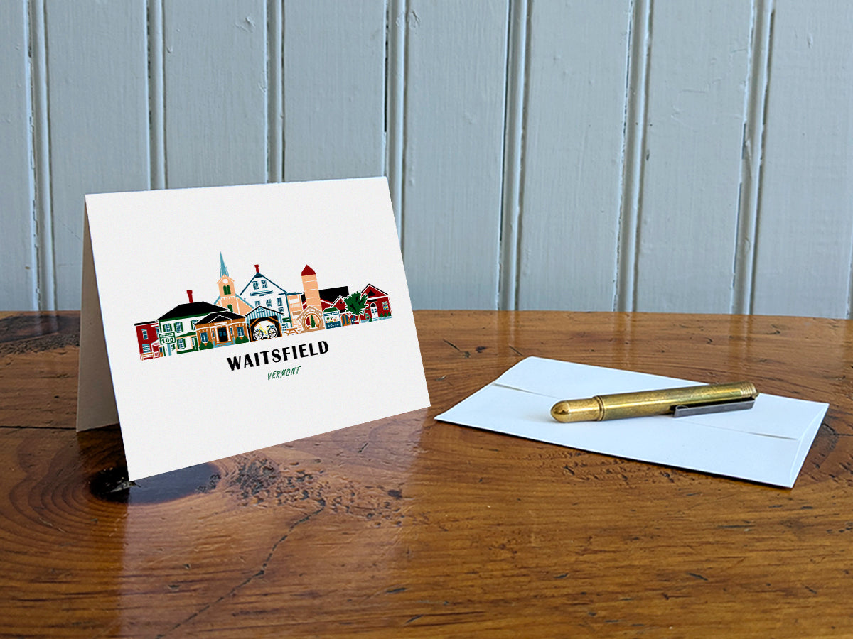 Greeting card with image of buildings arranged in a city skyline with the text Waitsfield Vermont on a table with gold pen and envelope.