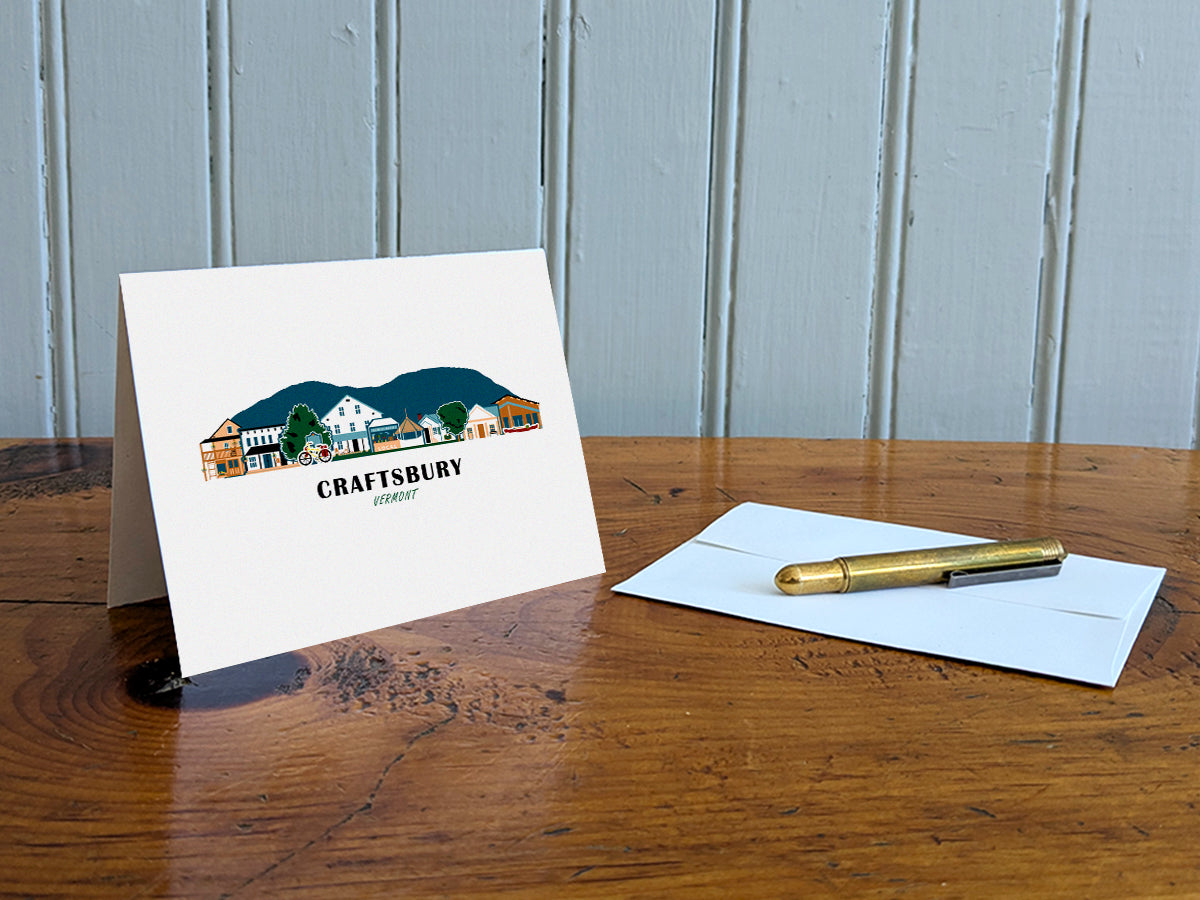 Greeting card with image of buildings arranged in a city skyline with the text Craftsbury Vermont on a table with gold pen and envelope.