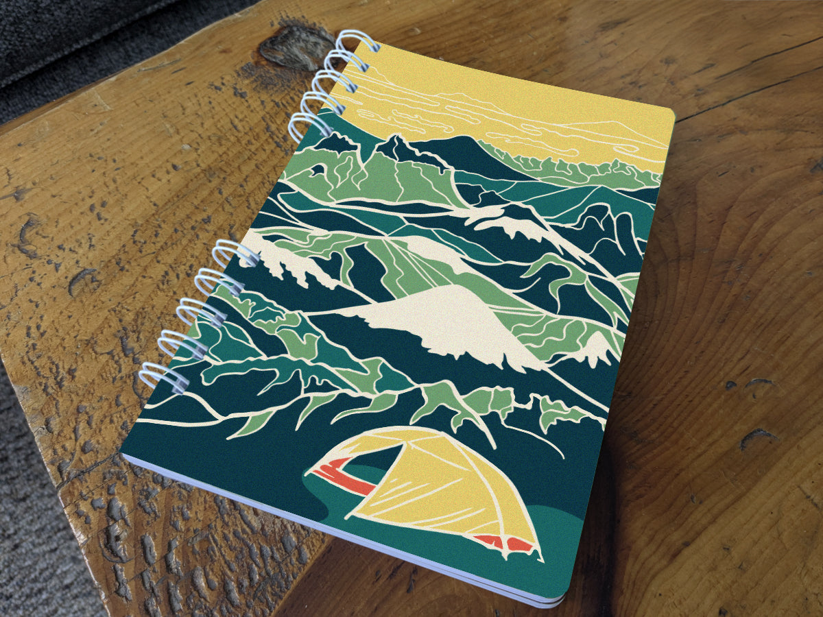 Notebook with illustration of a yellow tent among a background of blue and green mountains on a table.