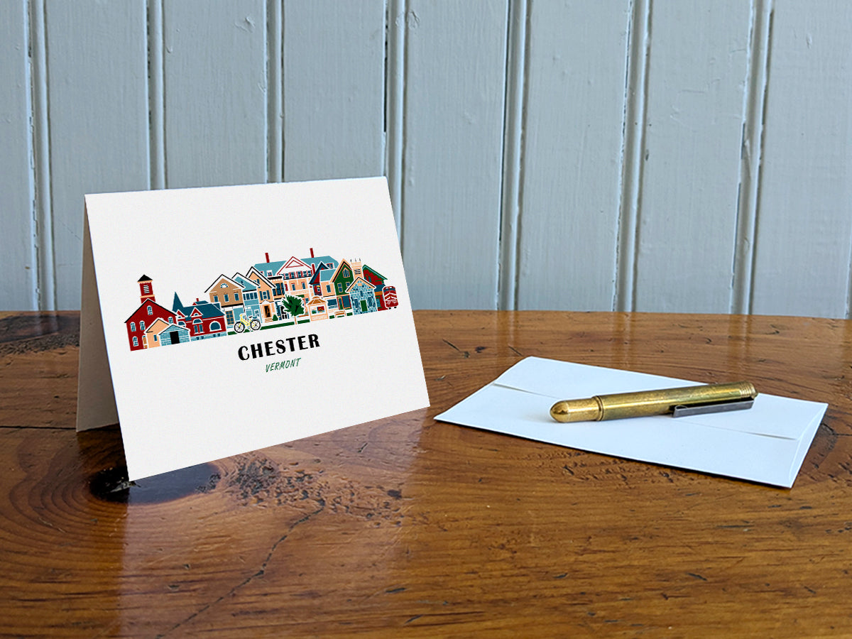 Greeting card with image of buildings arranged in a city skyline with the text Chester Vermont on a table with gold pen and envelope.