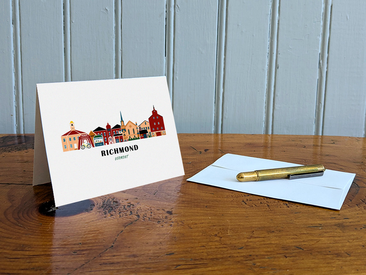 Greeting card with image of buildings arranged in a city skyline with the text Richmond Vermont on a table with gold pen and envelope.