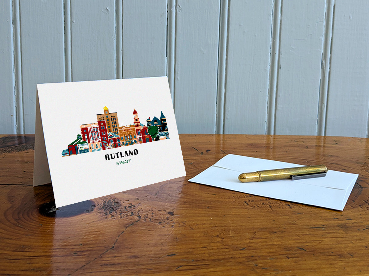 Greeting card with image of buildings arranged in a city skyline with the text Rutland Vermont on a table with gold pen and envelope.
