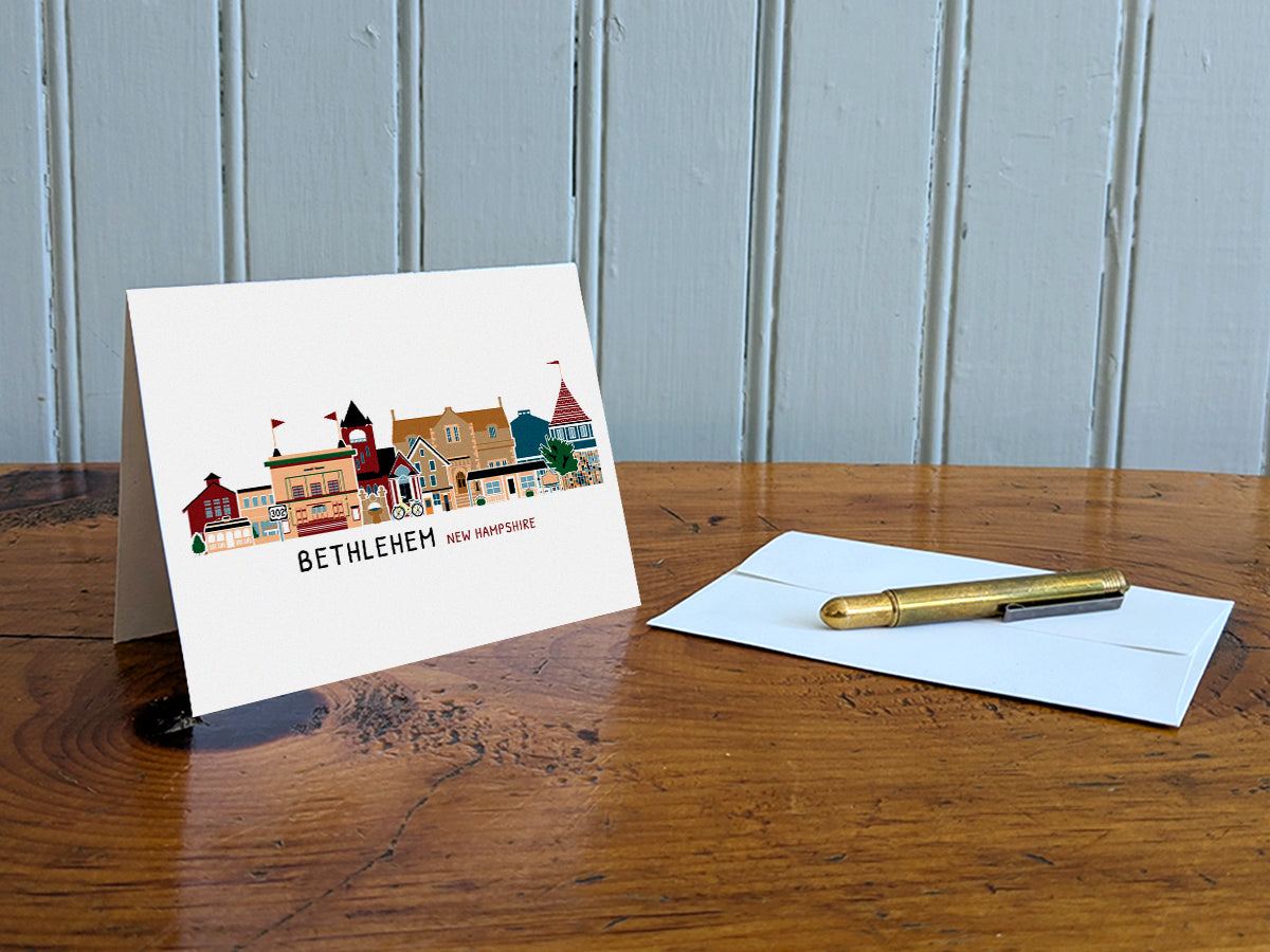Greeting card with illustration of Bethlehem, New Hampshire, on a wooden table with an envelope and pen.