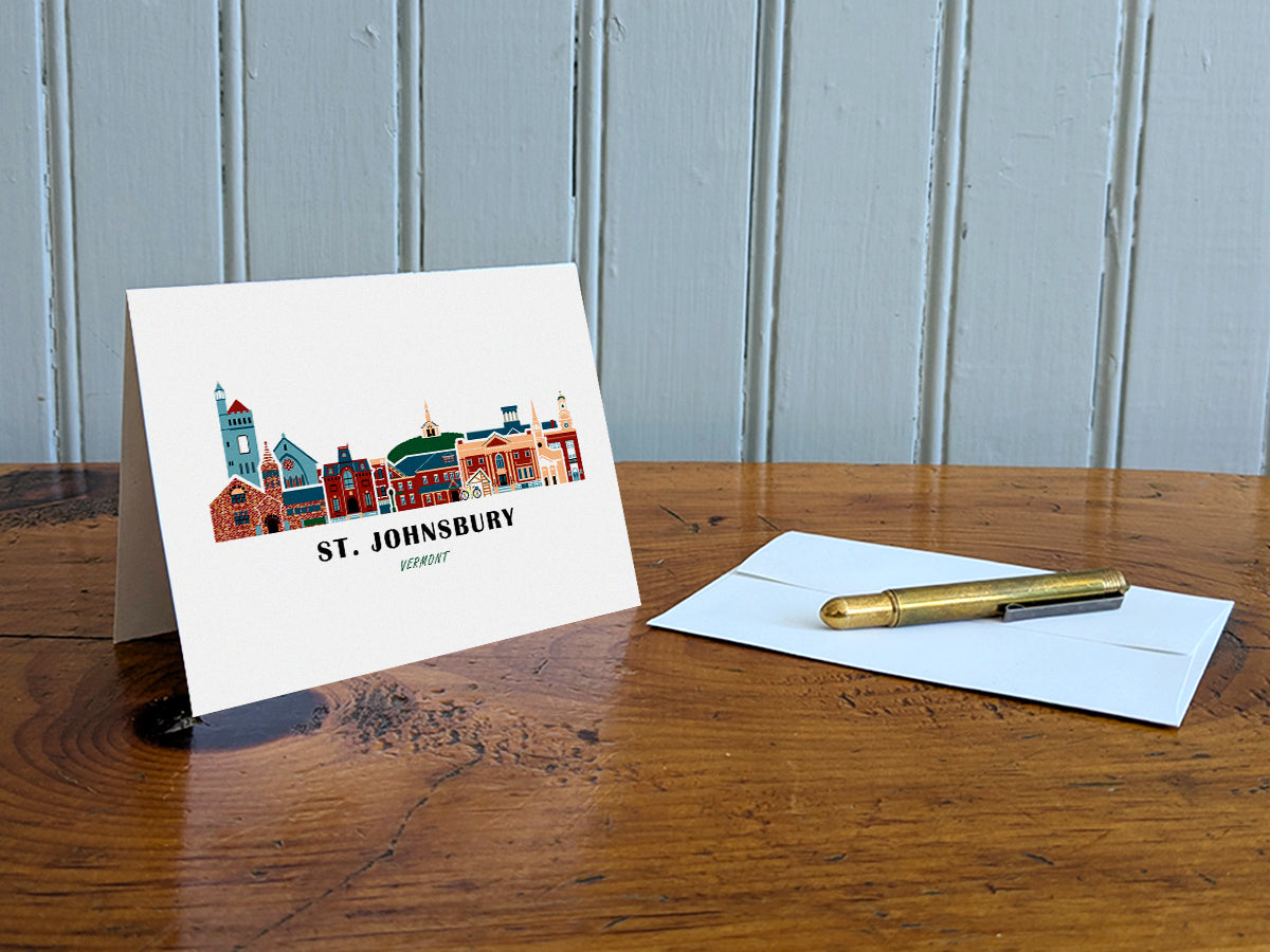 Greeting card with image of buildings arranged in a city skyline with the text St. Johnsbury Vermont on a table with gold pen and envelope.