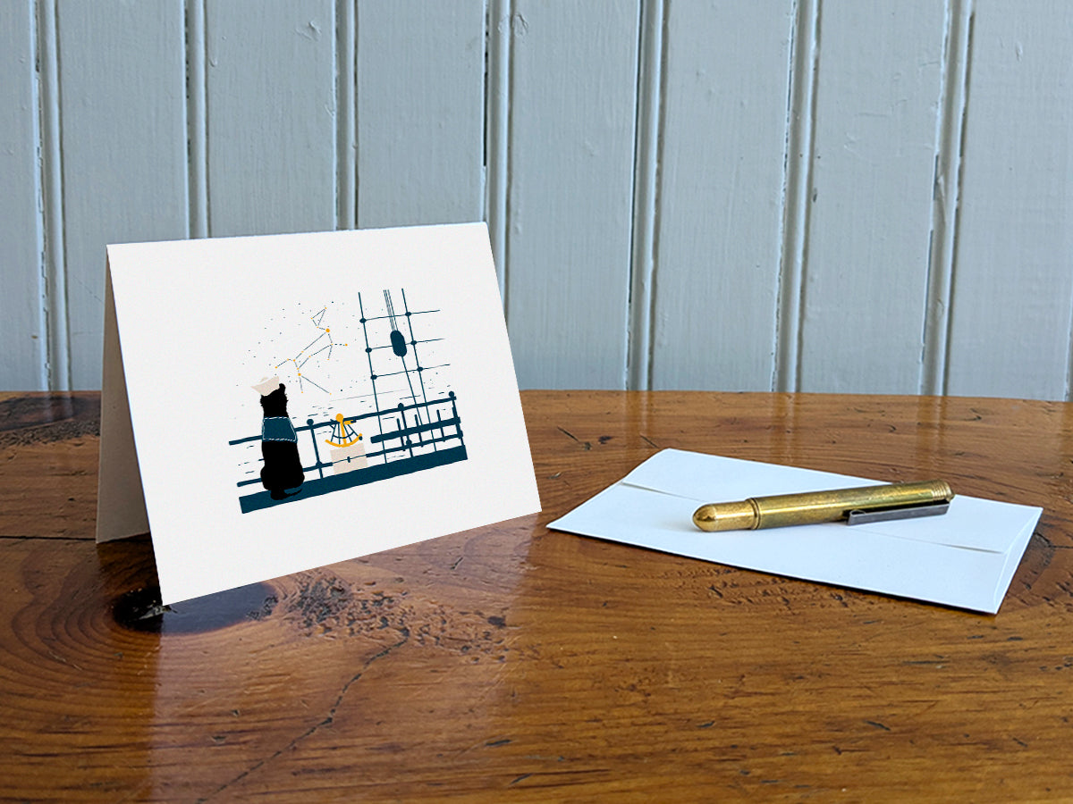 Greeting card with image of a dog with a sextant looking at the stars on a table with gold pen and envelope.