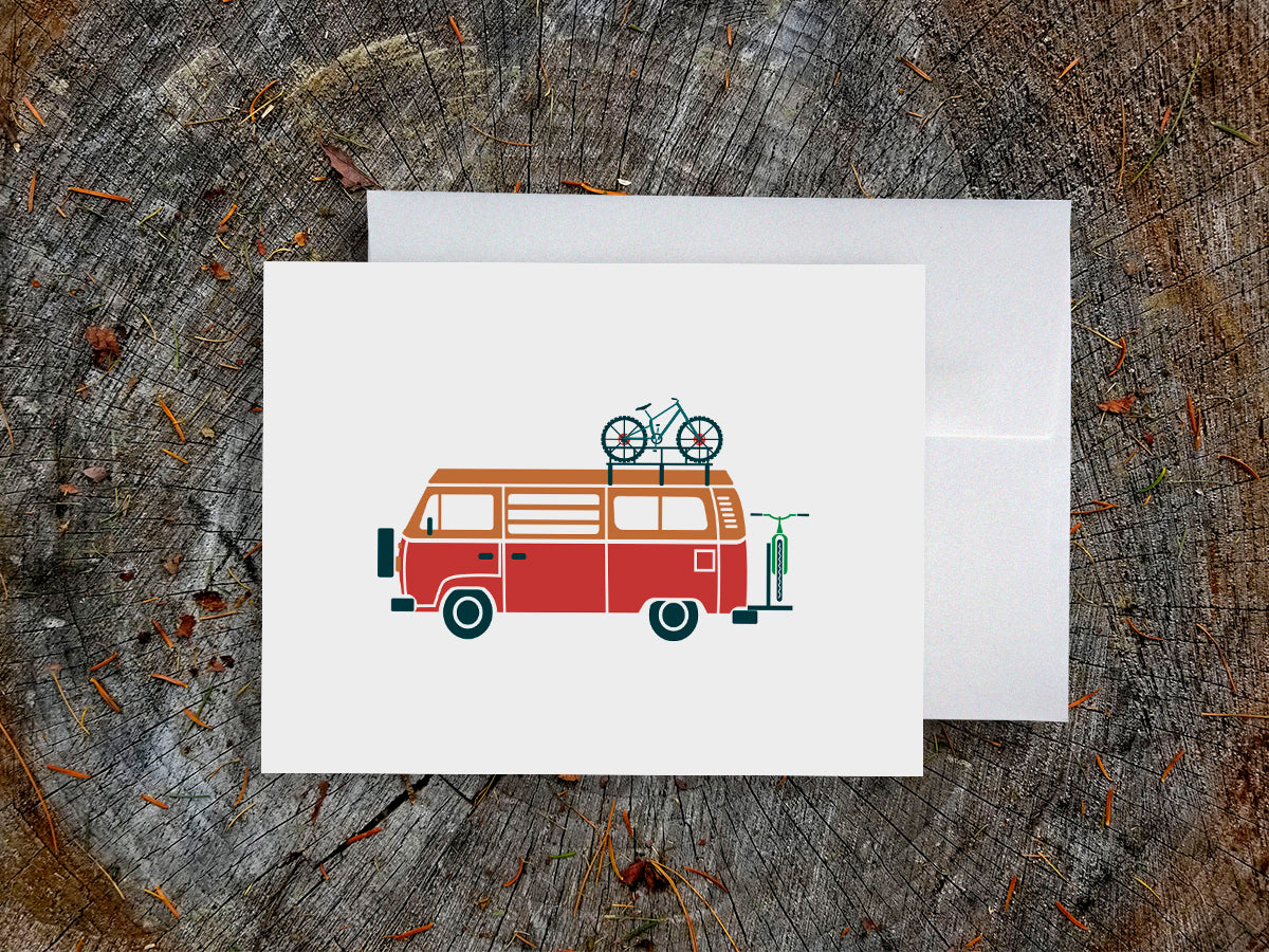 Greeting card with a red and orange camper van carrying two bikes with wood background