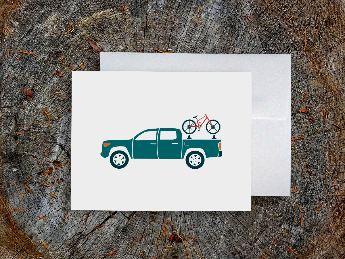 Greeting card with a blue truck carrying a bike with wood background
