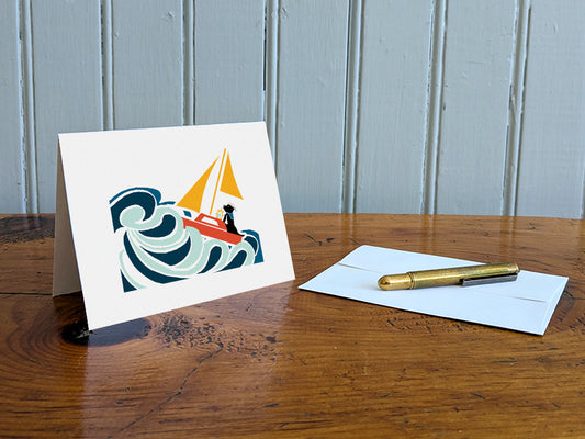 Greeting card with image of a dog in a sailboat on a table with gold pen and envelope.