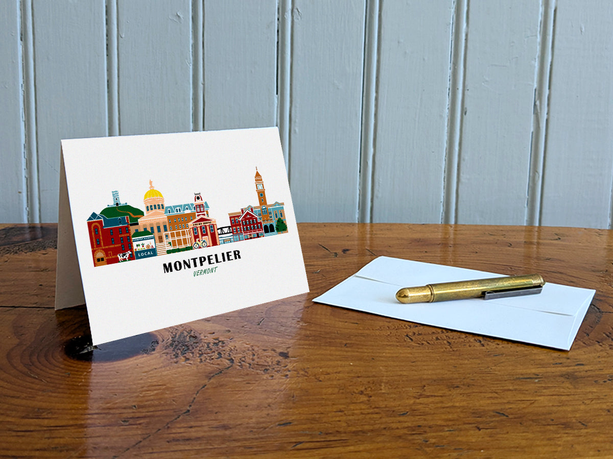 Greeting card with image of buildings arranged in a city skyline with the text Montpelier Vermont on a table with gold pen and envelope.
