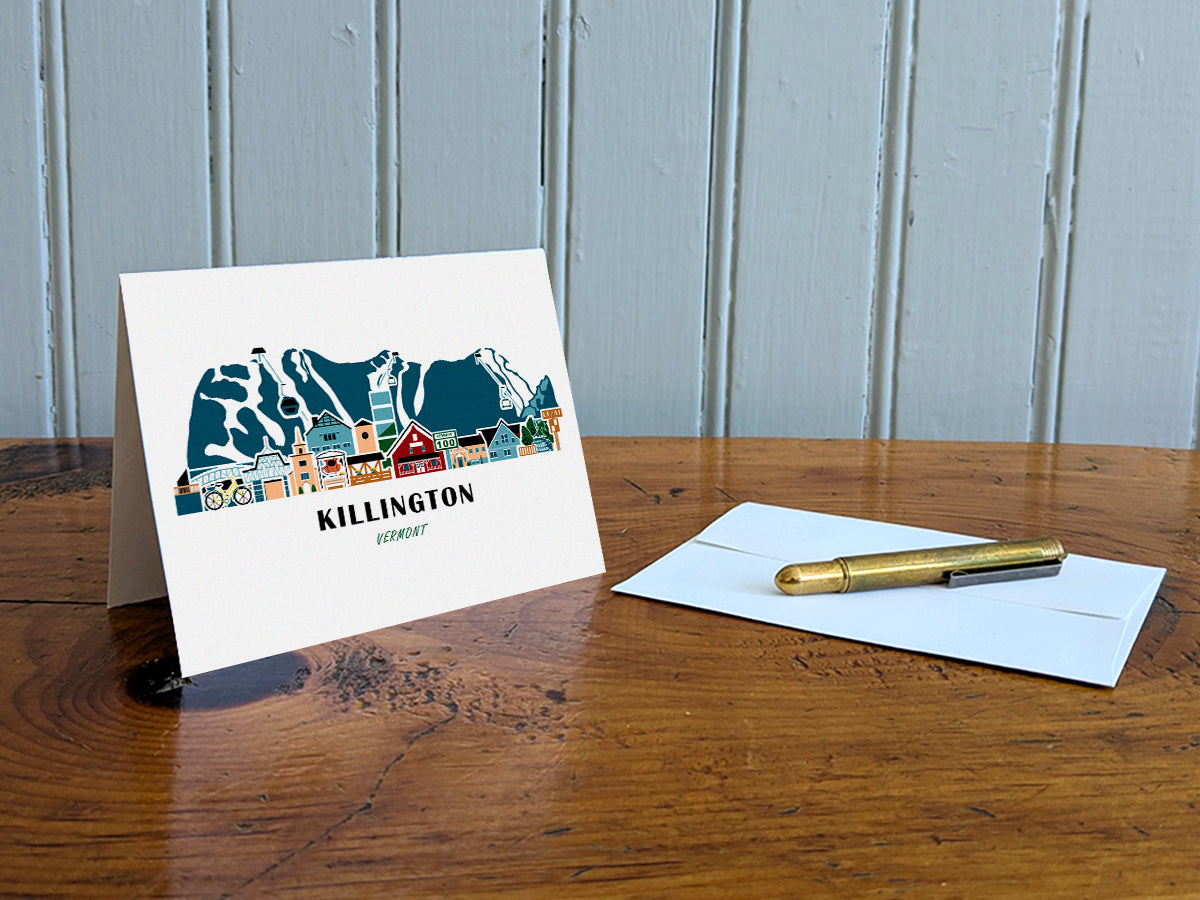 Greeting card with image of buildings arranged in a city skyline with the text Killington Vermont on a table with gold pen and envelope.