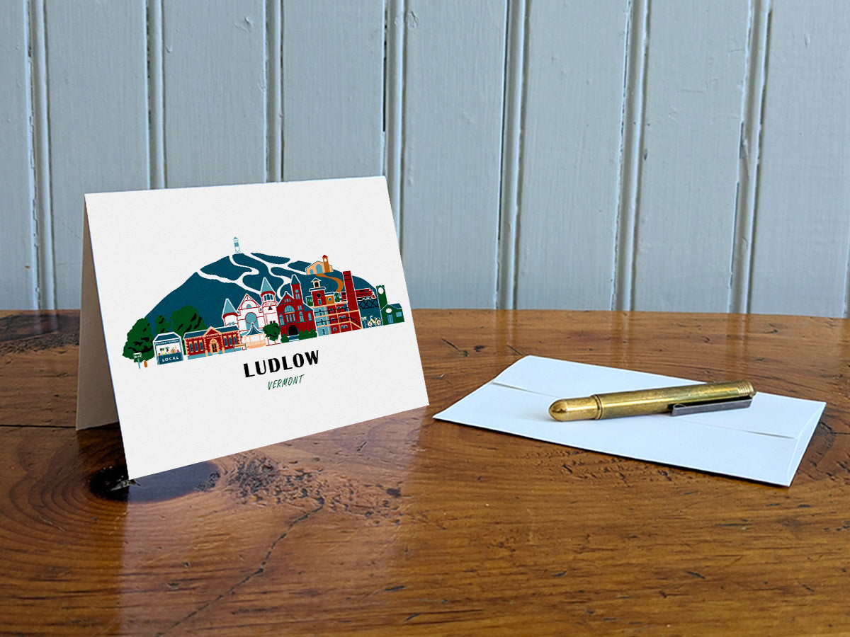 Greeting card with image of buildings arranged in a city skyline with the text Ludlow Vermont on a table with gold pen and envelope.