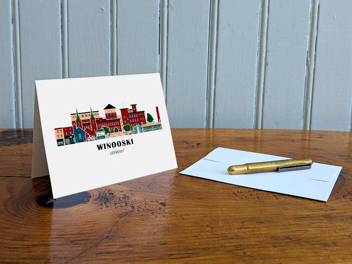 Greeting card with image of buildings arranged in a city skyline with the text Winooski Vermont on a table with gold pen and envelope.