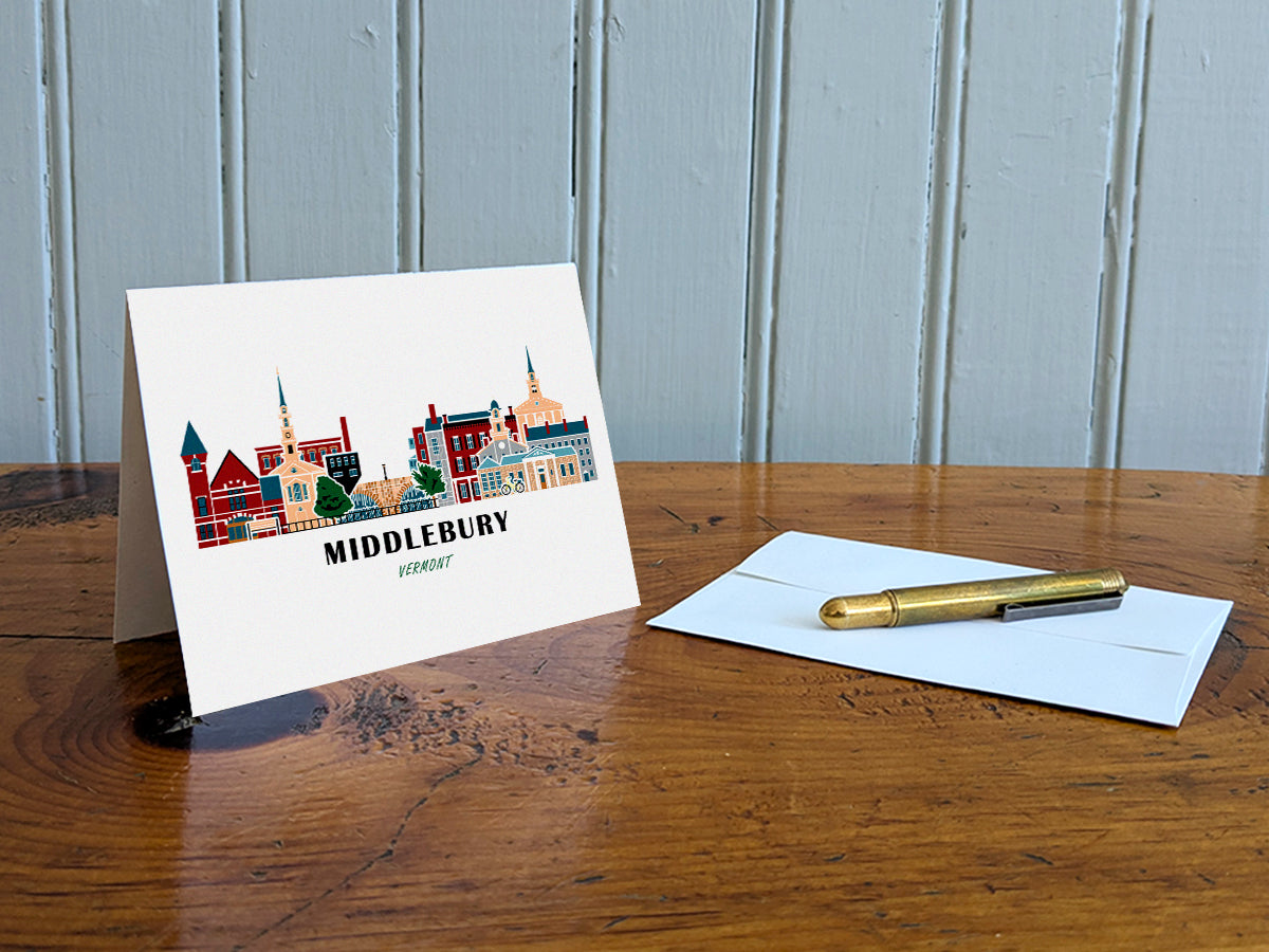 Greeting card with image of buildings arranged in a city skyline with the text Middlebury Vermont on a table with gold pen and envelope.