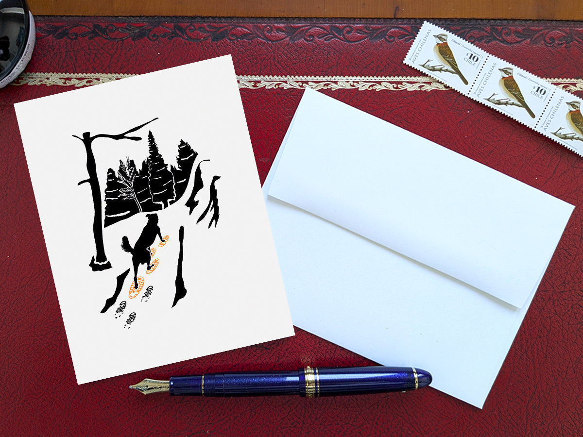 Greeting card with image of dog walking through forest wearing yellow snowshoes on a table with blue pen and envelope and stamps.