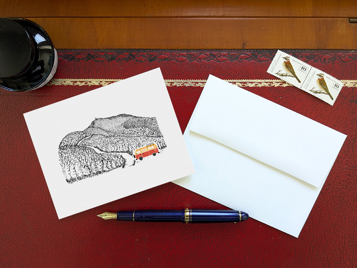 Greeting card with image of a mountain road and an orange and yellow campervan on a table with blue pen and envelope and stamps.
