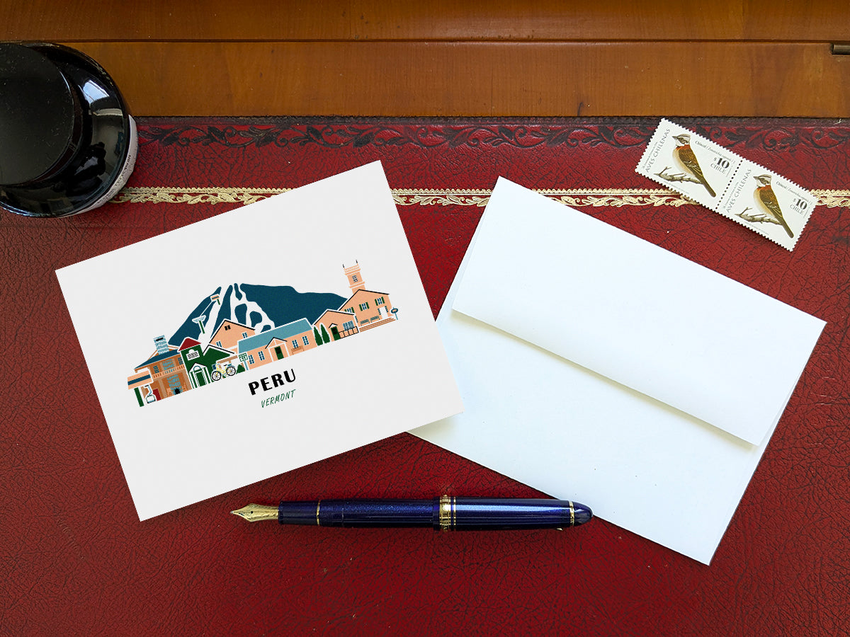 Greeting card with image of buildings arranged in a city skyline with the text Peru Vermont on a table with blue pen and envelope and stamps.