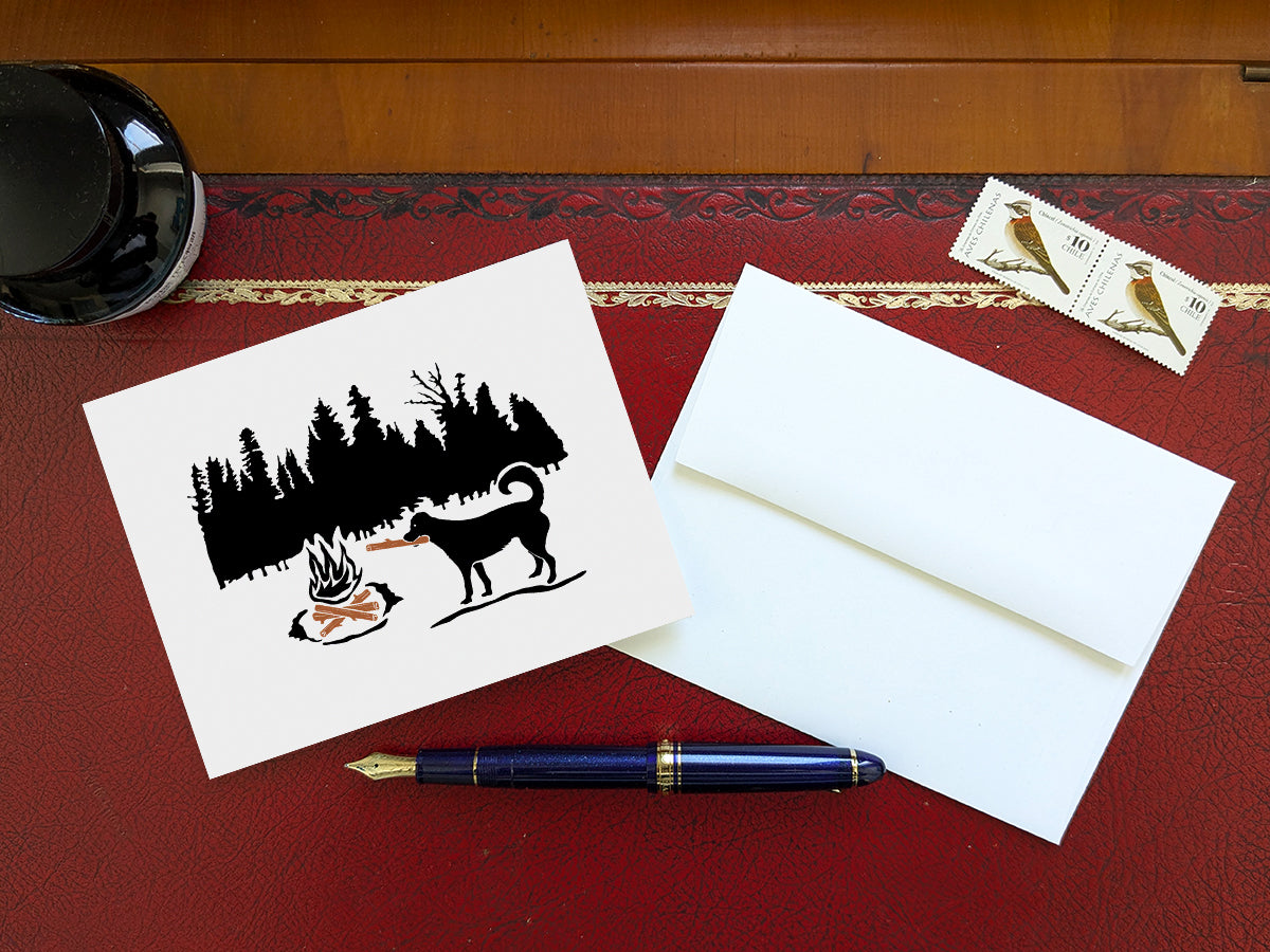Greeting card of black dog putting a log in a campfire with forest background on a table with blue pen and envelope and stamps.
