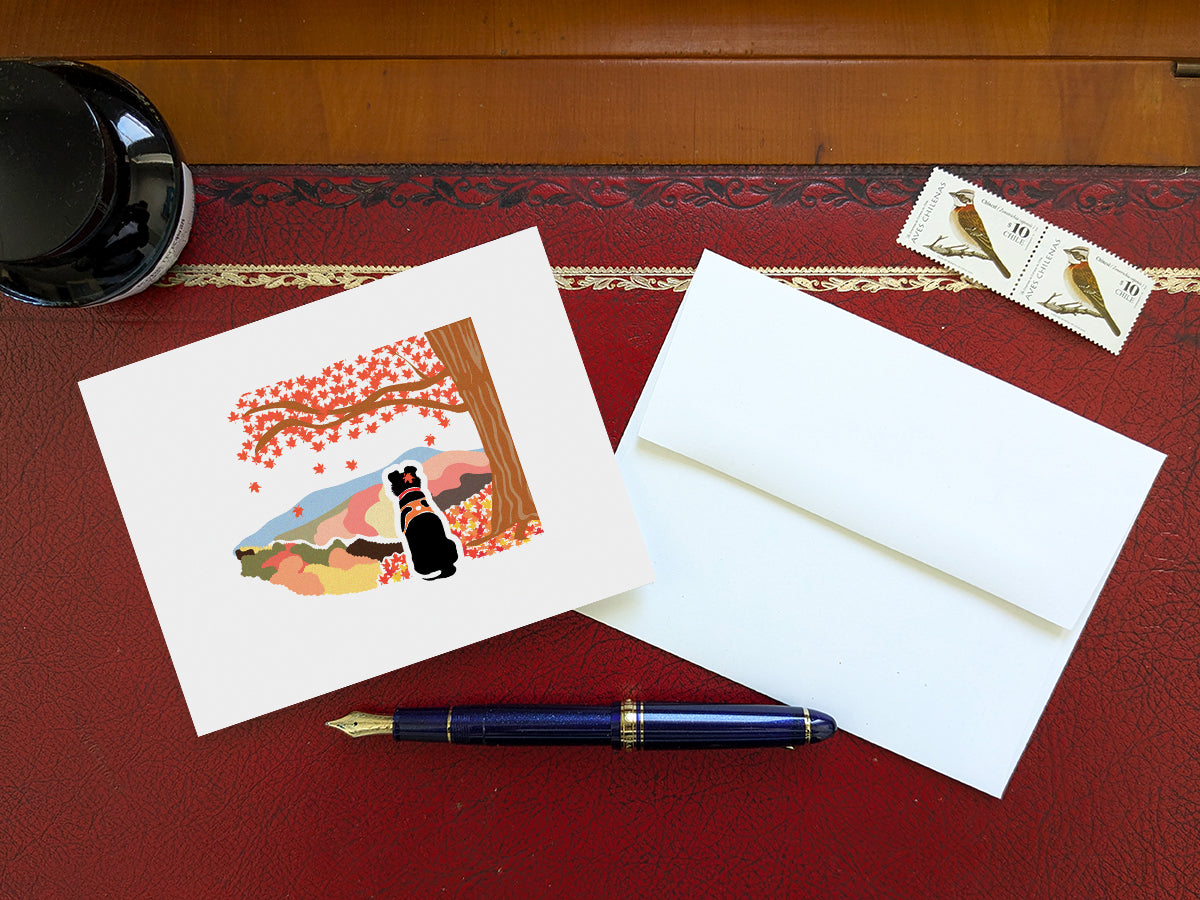 Greeting card with image of black dog underneath a maple tree with red fall foliage and mountains in the background on a table with a blue pen and envelope and stamps.