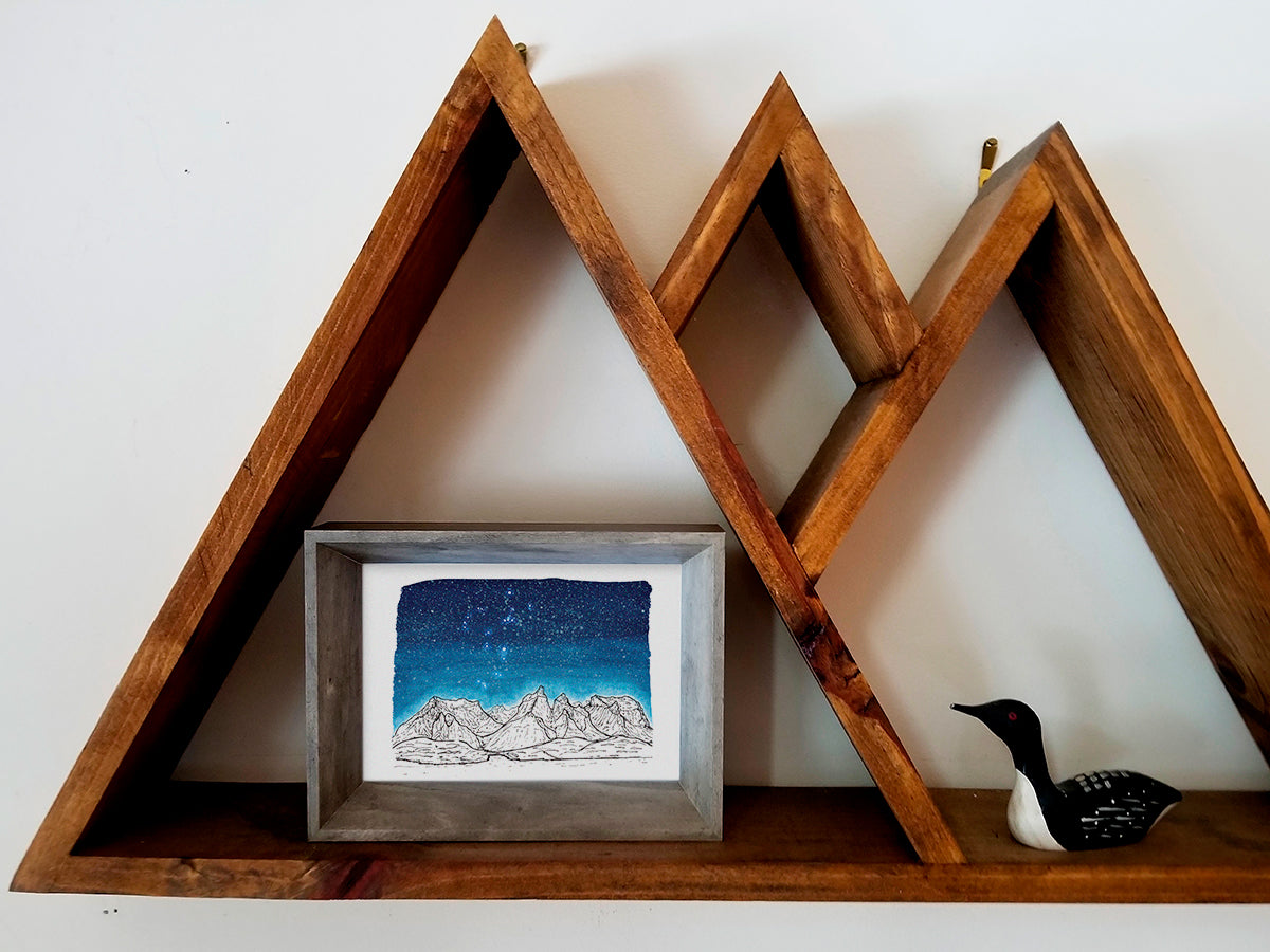 Framed art print on a wooden shelf of black and white mountains and a dark blue star filled sky