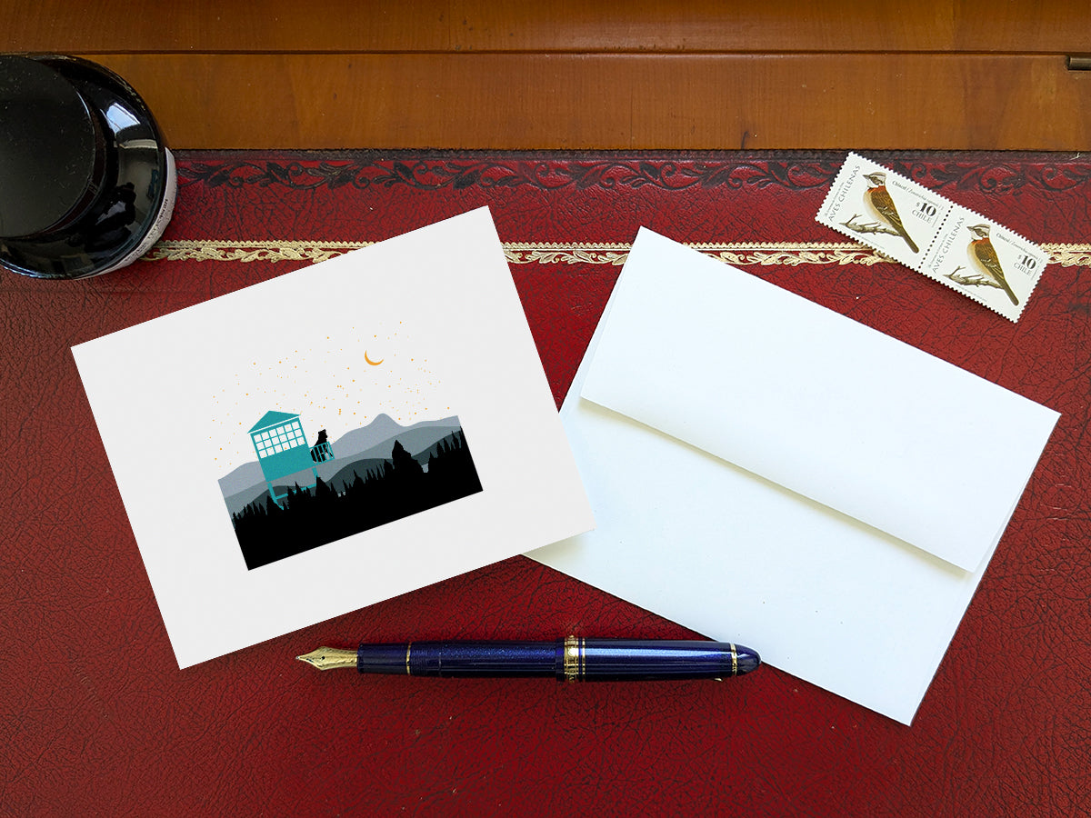 Greeting card of black dog sitting in a fire tower overlooking the mountains and stars on a table with a blue pen and envelope and stamps.