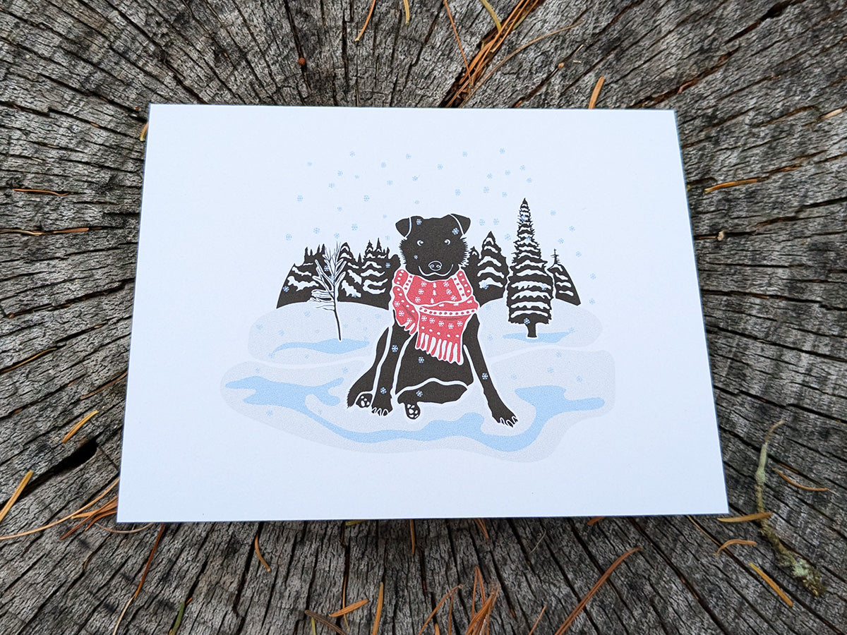 Greeting card with image of black dog with red scarf among a snowy winter landscape.
