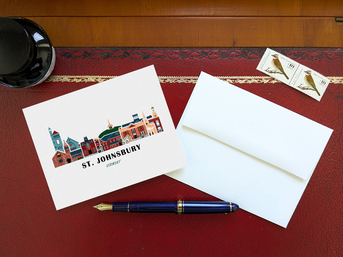 Greeting card with image of buildings arranged in a city skyline with the text St. Johnsbury Vermont on a table with blue pen and envelope and stamps.