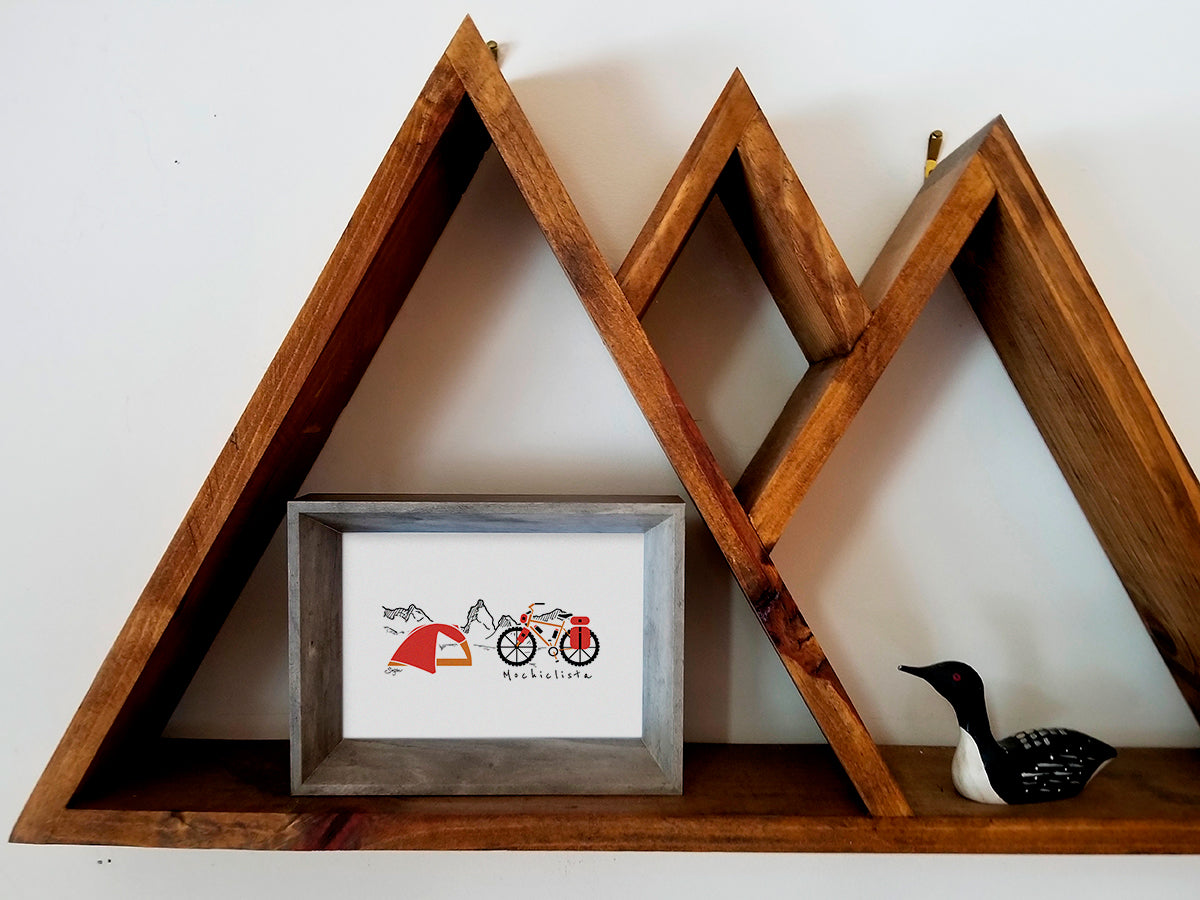 Framed art print on a wooden shelf of a red and orange bike and tent with mountains in the background and the text Mochiclista