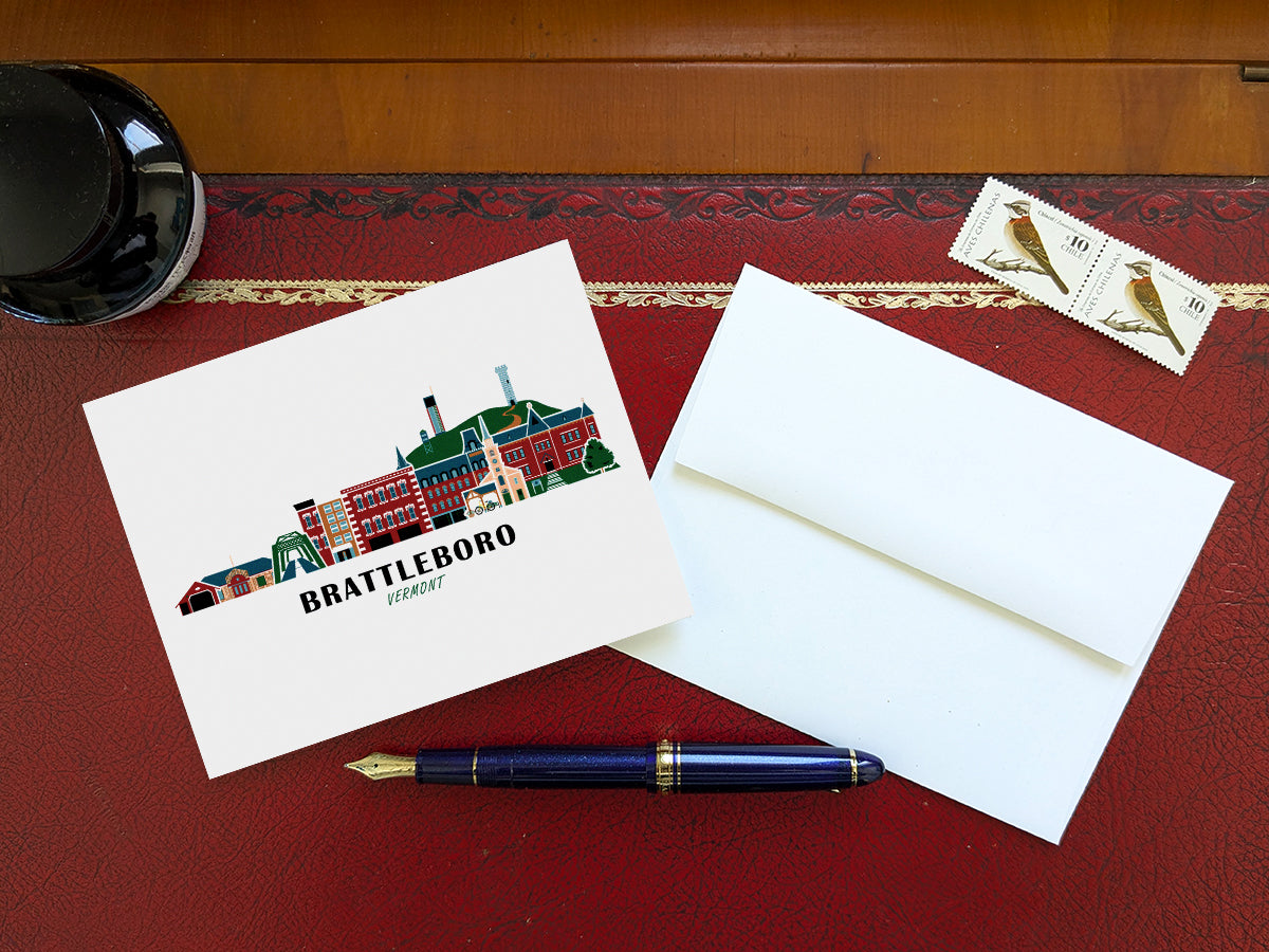 Greeting card with image of buildings arranged in a city skyline with the text Brattleboro Vermont on a table blue pen and envelope and stamps.
