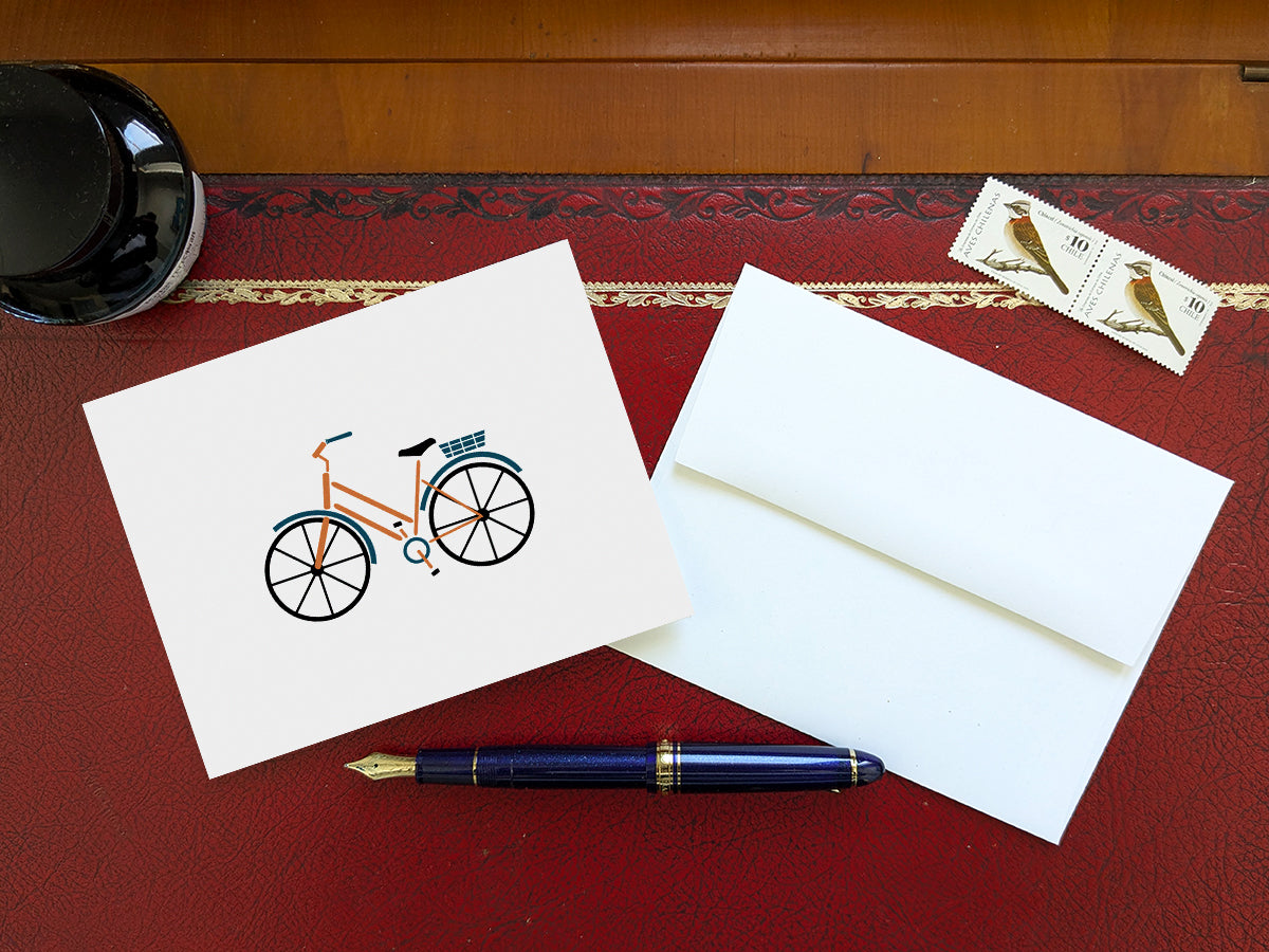 Greeting card with a yellow cruiser bike with basket on a table with a blue pen and envelope and stamps
