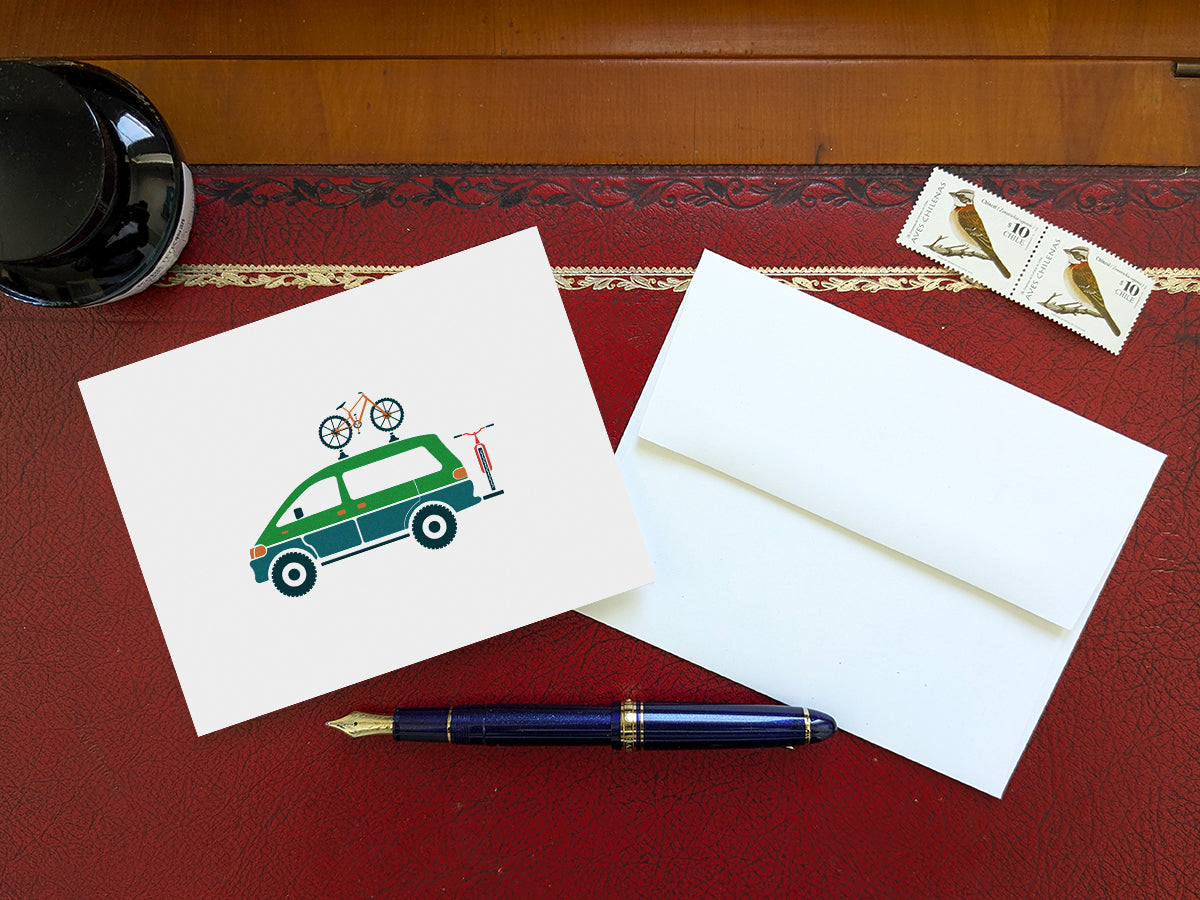 Greeting card with a blue and green van carrying two bikes on a table with a blue pen and envelope and stamps