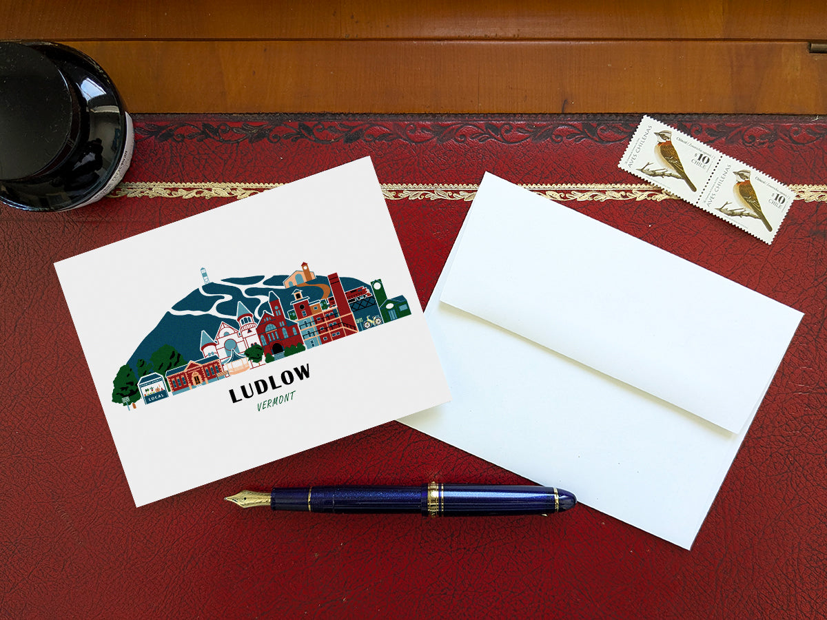 Greeting card with image of buildings arranged in a city skyline with the text Ludlow Vermont on a table with blue pen and envelope and stamps.