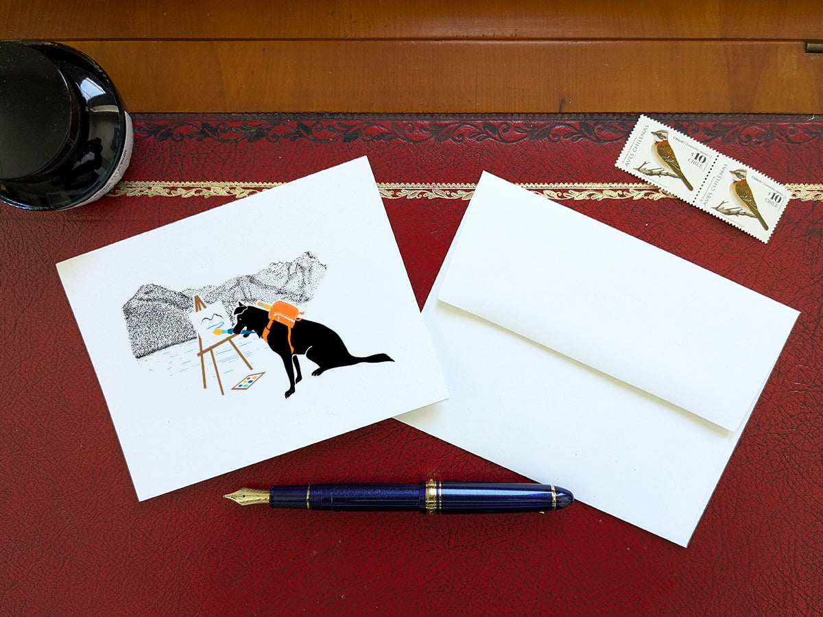 White card with drawing of dog painting in the mountains on a wooden surface with a pen, envelope, and stamp.