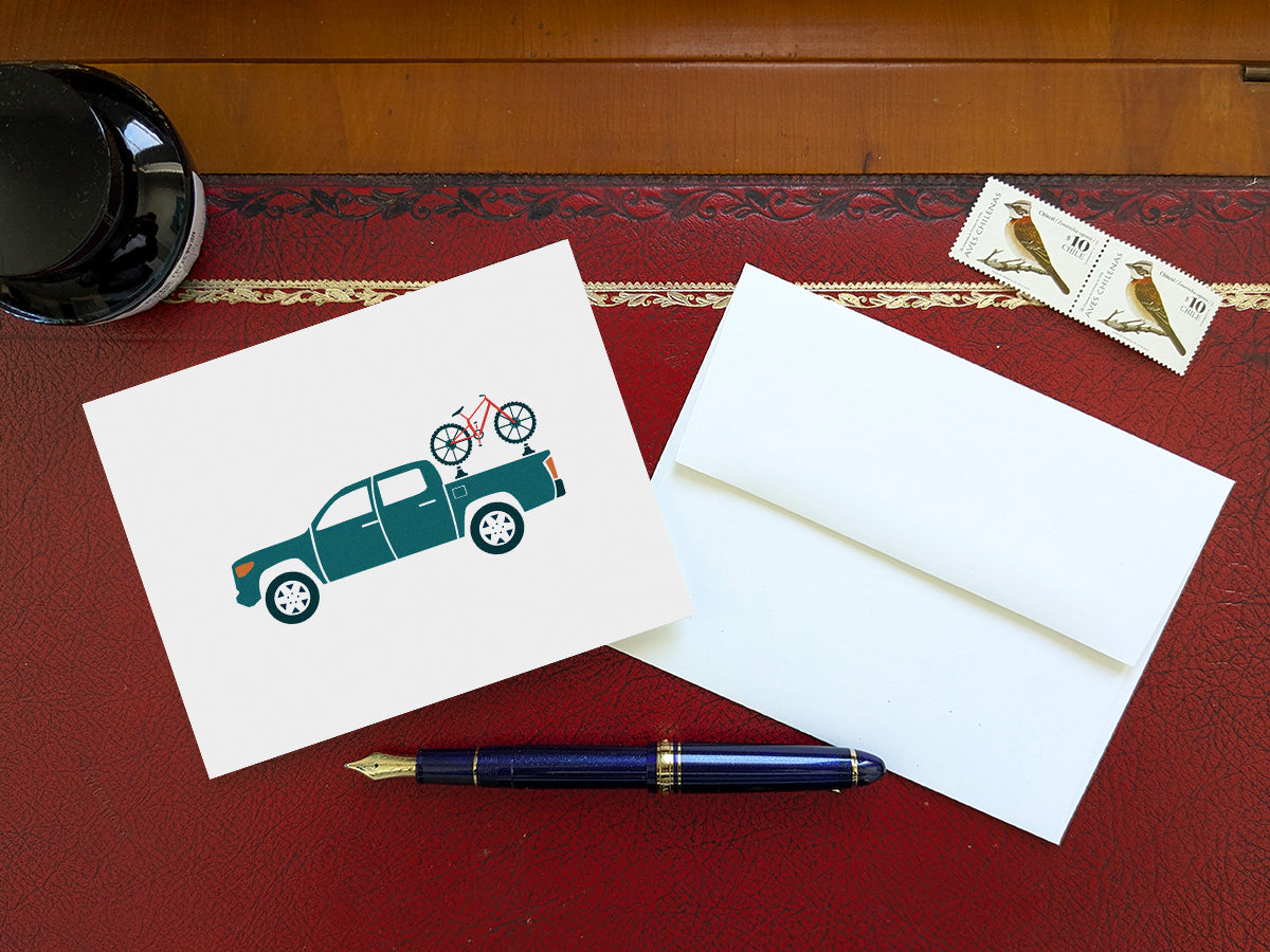 Greeting card of a blue truck carrying a bike on a table with a blue pen and envelope and stamps