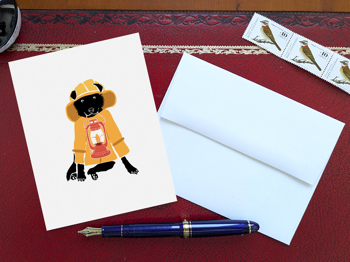 Greeting card with image of a dog with a yellow fisherman's jacket holding a lamp on a table with a blue pen and envelope and stamps.