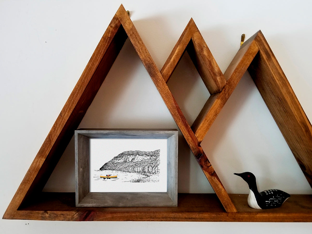 Framed art print placed on a wooden shelf of a dog paddling a yellow canoe with a mountain in the background