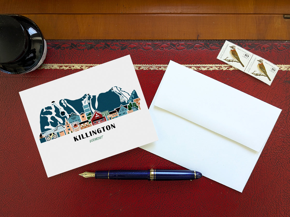 Greeting card with image of buildings arranged in a city skyline with the text Killington Vermont on a table with blue pen and envelope and stamps.