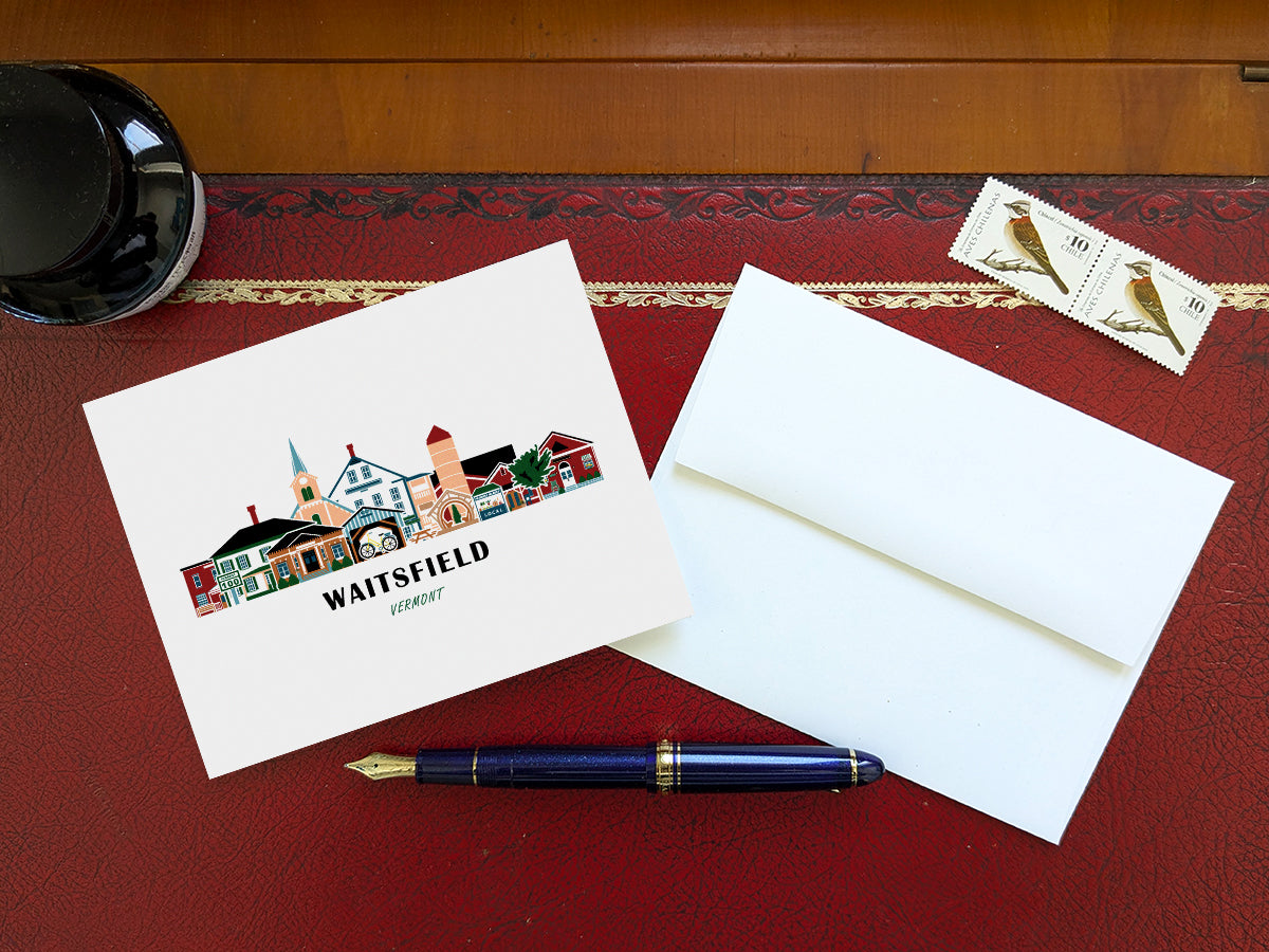 Greeting card with image of buildings arranged in a city skyline with the text Waitsfield Vermont on a table with blue pen and envelope and stamps.