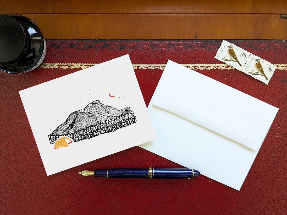 Greeting card with an image of a mountain and bright yellow tent under a starry sky on a table with blue pen and envelope and stamps.