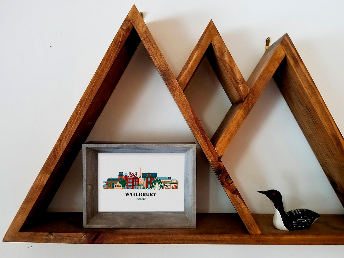 Framed art print on a wooden shelf of buildings arranged in a skyline with the text Waterbury Vermont